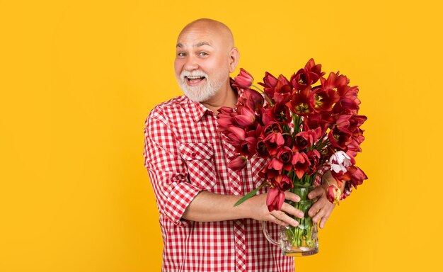 Fröhlicher alter Rentner mit Bart hält Frühlingstulpenblumen auf gelbem Hintergrund