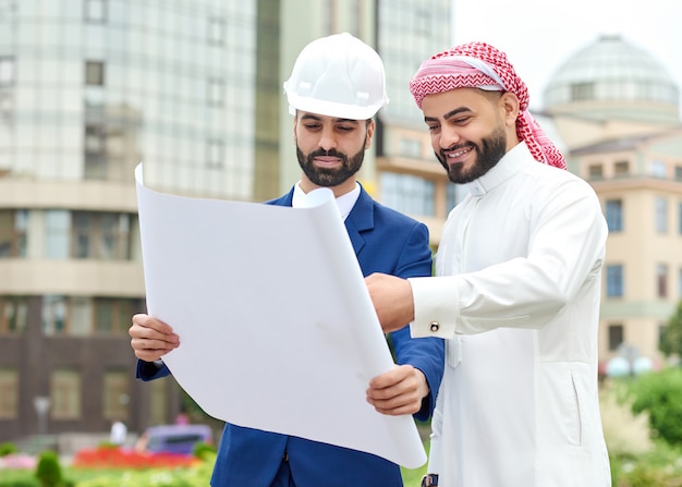 Fröhlicher arabischer Geschäftsmann, der mit seinem Architekten einige Pläne durchgeht