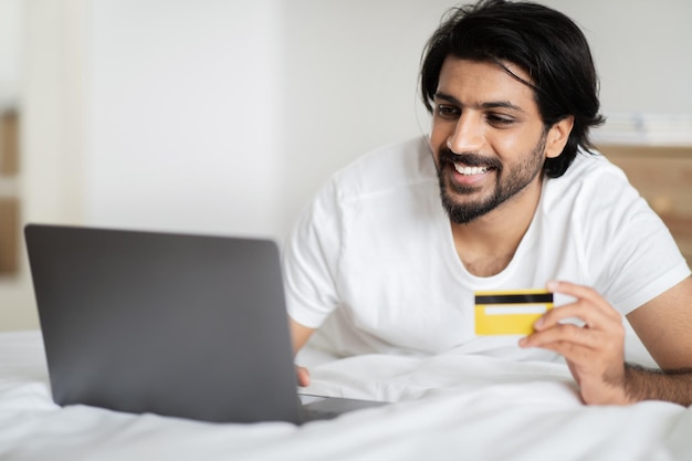 Fröhlicher arabischer Typ in weißem T-Shirt liegt mit Laptop und Kreditkarte auf dem Bett und genießt Online-Shopping