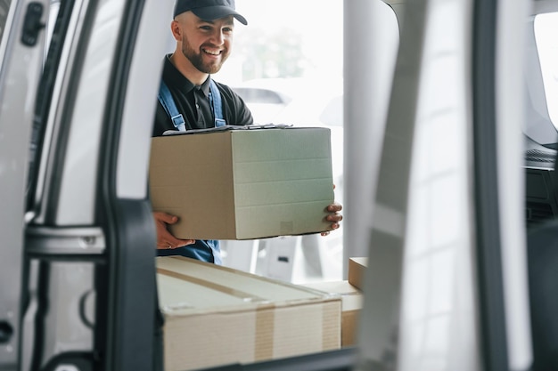 Fröhlicher Arbeiter Liefermann in Uniform ist drinnen mit Auto und mit Bestellung