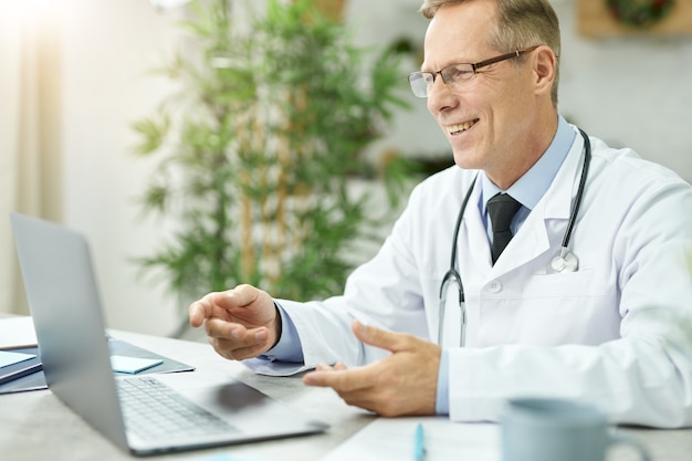 Fröhlicher Arzt, der mit Laptop am Tisch sitzt und lächelt, während er mit dem Patienten online spricht talking