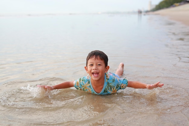 Fröhlicher asiatischer Junge spielt und liegt gerne am tropischen Sandstrand bei Sonnenaufgang Entzückendes kleines Kind, das Spaß in den Sommerferien hat