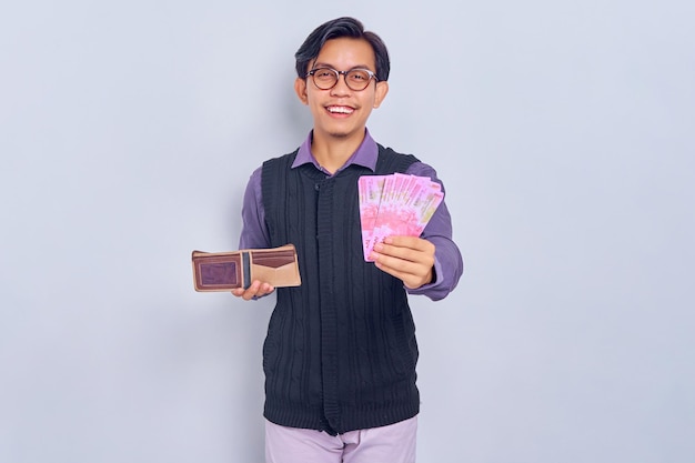 Fröhlicher asiatischer junger Mann mit Hemd, der Brieftasche und Rupiah-Bargeldbanknote isoliert auf weißem Hintergrund hält