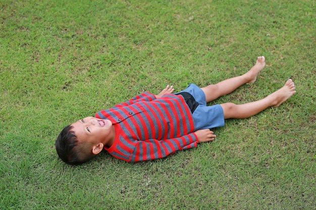 Fröhlicher asiatischer kleiner Junge, der sich auf grünem Rasen hinlegt Kid liegt auf Gras und blickt auf den Himmel