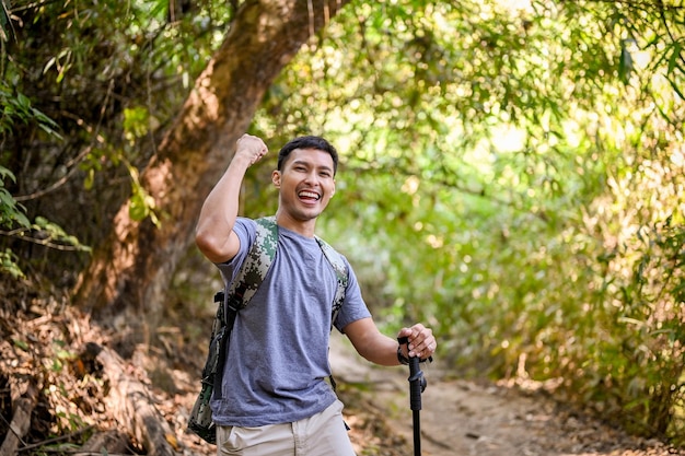 Fröhlicher asiatischer männlicher Reisender mit Trekkingausrüstung und Rucksackhand-up-Triumphgeste feiert