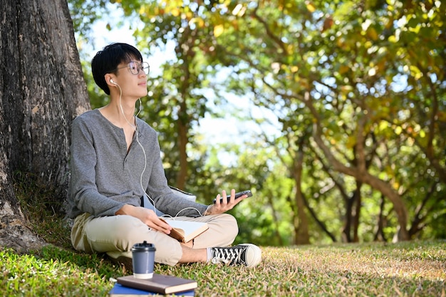 Fröhlicher asiatischer männlicher Student, der Musik hört, während er sich unter dem Baum im Park entspannt