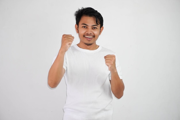 fröhlicher asiatischer Mann hebt die Hände hoch, trägt ein T-Shirt, gewinnende Geste, drückt die Fäuste zusammen, fühlt sich glücklich
