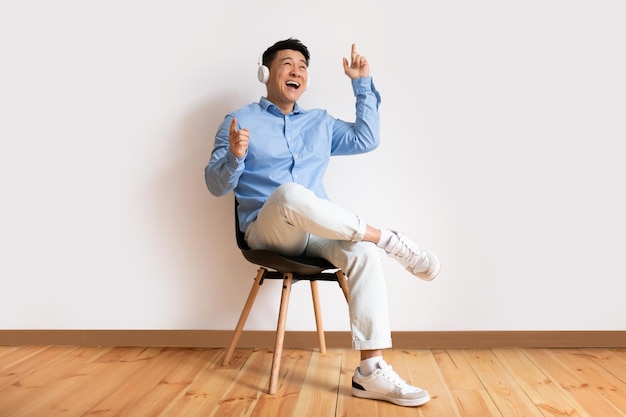 Fröhlicher asiatischer reifer Mann, der mit Kopfhörern im Stuhl sitzt, Musik hört und Kopierraum tanzt