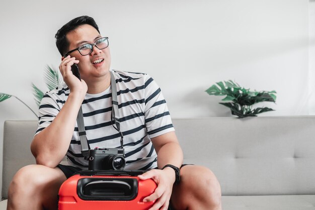 Fröhlicher asiatischer Reisender mit Brille und Kamera, der auf der Couch sitzt, während er im Wohnzimmer mit dem Handy telefoniert Reise- und Urlaubskonzept