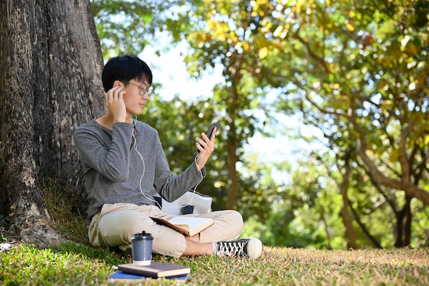 Fröhlicher asiatischer Student, der Musik hört, während er sich unter dem Baum im Park entspannt