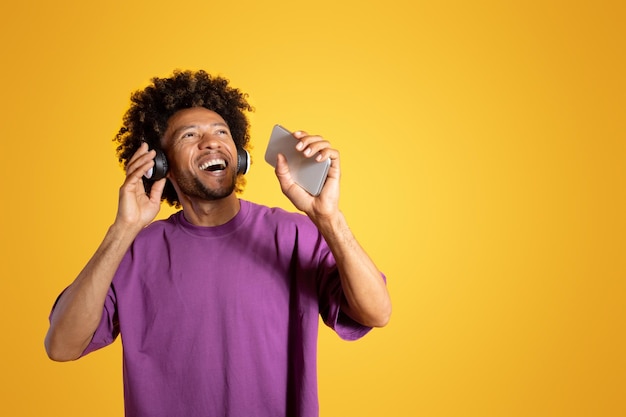 Fröhlicher aufgeregter erwachsener afroamerikanischer lockiger Mann in Kopfhörern mit offenem Mund singt am Smartphone