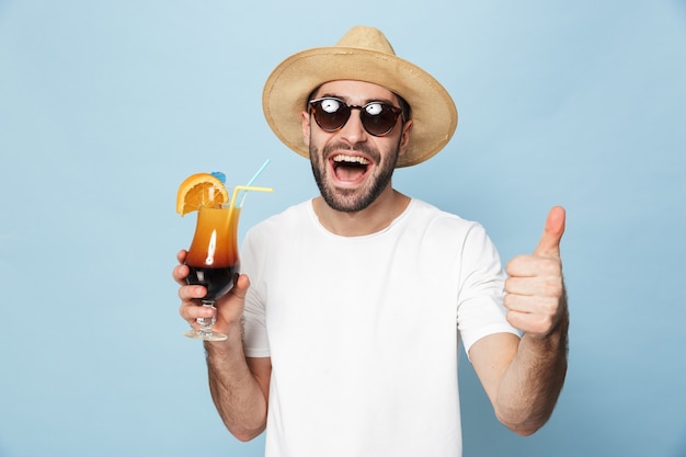 Fröhlicher aufgeregter Mann mit leerem T-Shirt, der isoliert über blauer Wand steht, Cocktail trinkt, Spaß hat, Daumen hoch