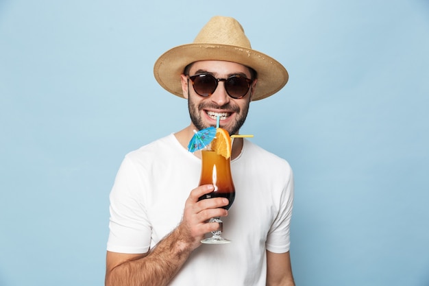 Fröhlicher aufgeregter Mann mit leerem T-Shirt, der isoliert über blauer Wand steht, Cocktail trinkt, Spaß hat