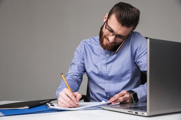 Fröhlicher, bärtiger, eleganter Mann mit Brille, der mit dem Smartphone spricht und etwas schreibt, während er im Büro am Tisch sitzt