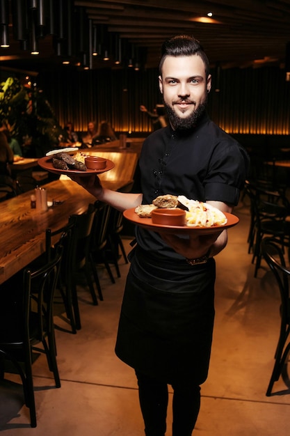 Fröhlicher bärtiger Kellner mit Kebab, Grill und Beilage in einem georgischen Restaurant. Ein anspruchsvoller Kellner in schwarzer Kleidung, mit Bart und einer Portion Döner.