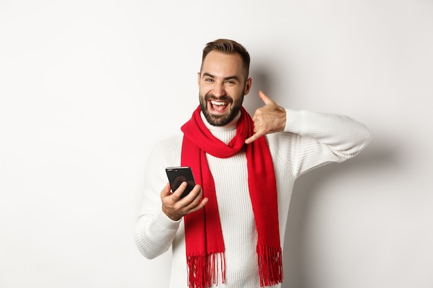 Fröhlicher bärtiger Kerl, der Smartphone hält, Telefonschild zeigt, darum bittet, ihn anzurufen, in Weihnachtspullover und Schal stehend, weißer Hintergrund