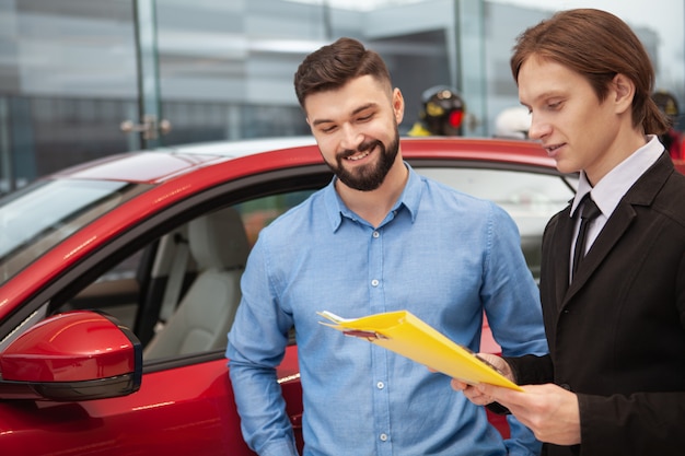 Fröhlicher bärtiger Mann, der mit Autohändler spricht, neues Auto im Autohaus kauft, Raum kopiert