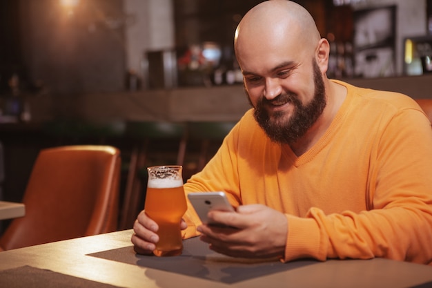 Fröhlicher bärtiger Mann, der sein Smartphone benutzt, während er Bier in der Kneipe trinkt