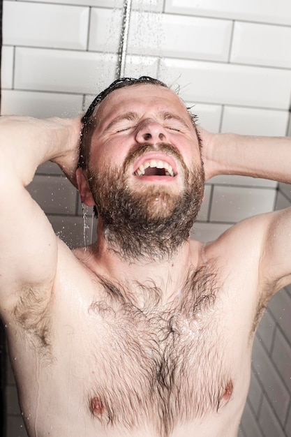 Fröhlicher bärtiger Mann, der seine Haare in der Dusche unter fließendem Wasser wäscht und glücklich lächelt