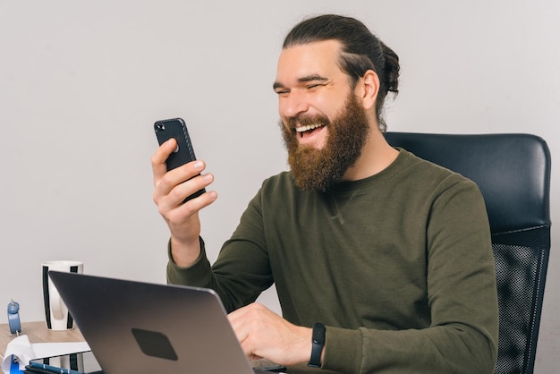 Fröhlicher bärtiger Mann lacht, während er sein Telefon und seinen Laptop benutzt.