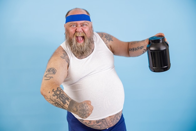 Foto fröhlicher bärtiger mann mit übergewicht hält eine schwarze flasche proteinpräparat auf hellblauem hintergrund