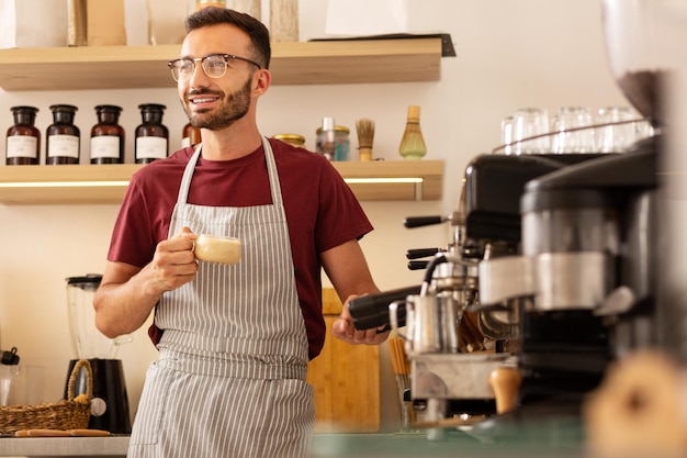 Fröhlicher Barista, der neben einer Kaffeemaschine mit einer Tasse Kaffee steht