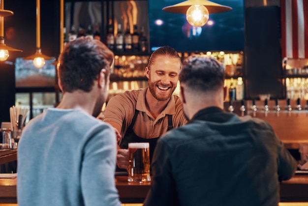 Fröhlicher Barkeeper, der jungen Männern Bier serviert, während er an der Bartheke in der Kneipe steht?