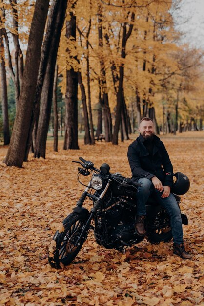 Fröhlicher Biker posiert, um Pause zu machen, nachdem er in der Nähe von schwarzem Motorrad gefahren ist, hält Schutzhelm, gelbe Bäume und Blätter herum, genießt gutes Wetter Herbstsaison Urbaner Lebensstil Außenaufnahme