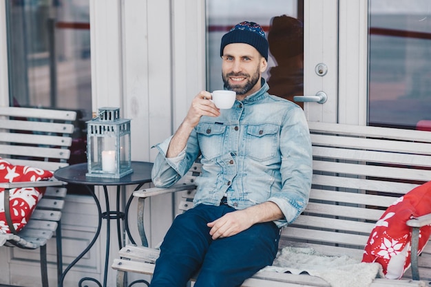 Fröhlicher blauäugiger Mann mit positivem Blick sitzt vor gemütlichem Interieur in der Terrassencafeteria genießt aromatischen Kaffee hat Freizeit nach der Arbeit Schöner Mann mit Bart sieht freudig in die Kamera