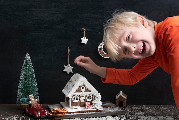 Fröhlicher blonder Junge neben Lebkuchenhaus Weihnachtshandwerk für Schule und Kindergarten