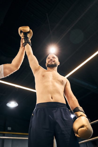 Fröhlicher Boxer in schwarzen Shorts, der nach dem Sieg mit der Hand über dem Kopf in einem hellen Ring bleibt