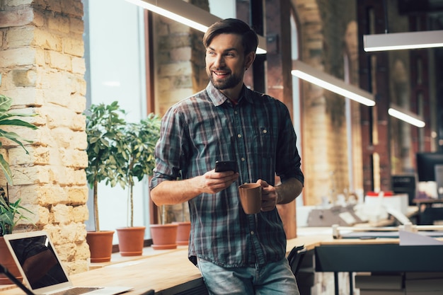 Fröhlicher Büroangestellter, der mit einer Tasse und einem Smartphone steht und lächelt, während er wegschaut