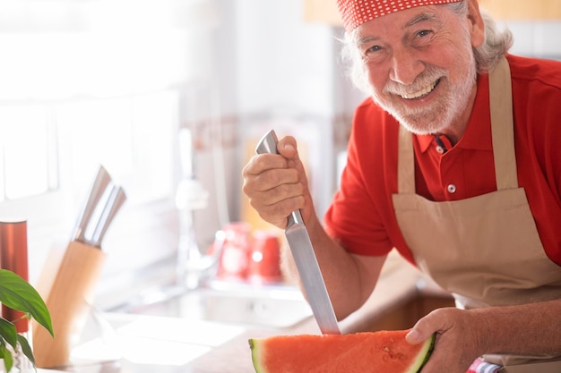 Fröhlicher Chefkoch in der Küche mit rotem Bandana beim Schneiden von Wassermelonen mit einem langen Messer - lächelnde aktive Rentner und lustiges Konzept