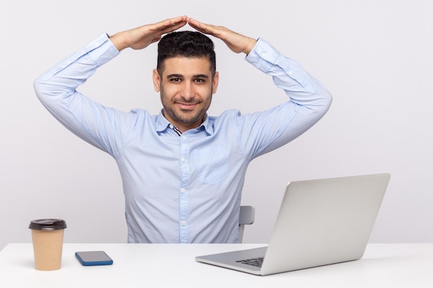 Fröhlicher, eleganter Angestellter, Versicherungsagent, der mit Laptop am Büroarbeitsplatz sitzt, die Geste des Hausdachs über dem Kopf hält und in die Kamera lächelt. Indoor-Studioaufnahme isoliert auf weißem Hintergrund