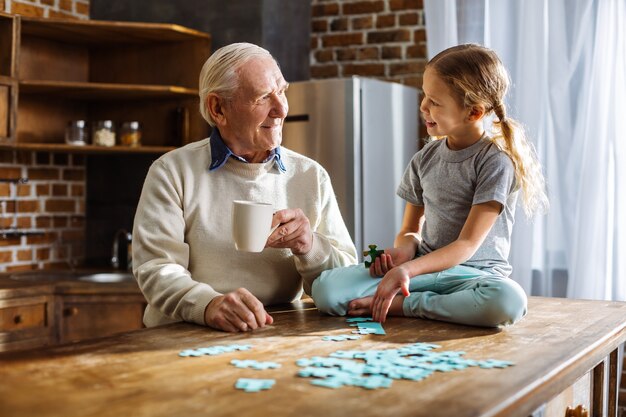 Fröhlicher elerly Mann, der Puzzles mit seiner Enkelin zusammenbaut, während er zu Hause ruht