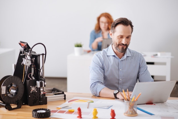 Fröhlicher entzückter gutaussehender Mann, der am Laptop arbeitet und ein neues 3D-Design entwickelt, während er am Tisch sitzt