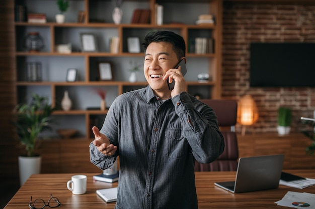 Fröhlicher, erfolgreicher, gutaussehender, reifer chinesischer Mann, der telefonisch mit dem Kunden im Innenbereich des Heimbüros telefoniert