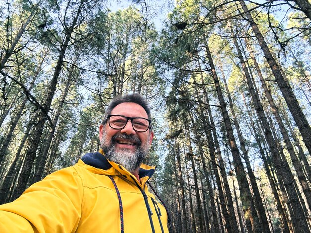 Fröhlicher, erwachsener, glücklicher Mann macht ein Selfie im Wald, genießt Freizeitaktivitäten im nationalen Waldpark allein, Abenteuer und Lebensstil, Menschen, hohe Bäume im Hintergrund, Nachhaltigkeit