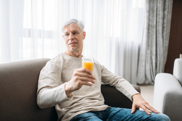 Fröhlicher erwachsener mann, der zu hause auf der couch sitzt und frischen saft trinkt. reife männliche person in jeans entspannen im sessel