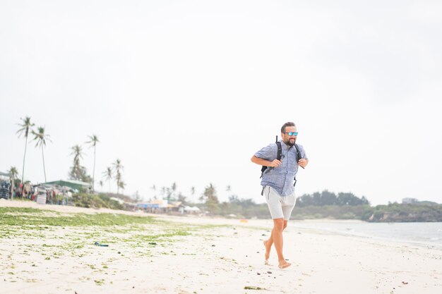 Fröhlicher erwachsener Mann mit Tasche, die Spaß am tropischen Strand hat