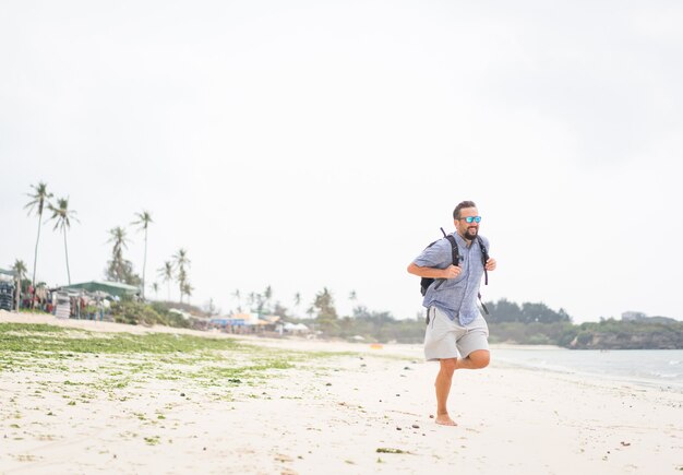 Fröhlicher erwachsener Mann mit Tasche, die Spaß am tropischen Strand hat