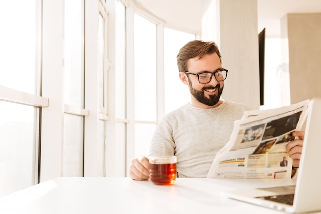 Fröhlicher europäischer bärtiger Mann, der Brille trägt, die am Tisch sitzt und Tee aus Glas trinkt, während Zeitung in Moring liest