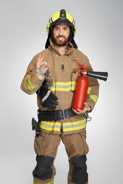 Fröhlicher Feuerwehrmann mit Feuerlöscher zeigt drinnen ein Okay-Schild, Vorderansicht eines bärtigen Feuerwehrmanns