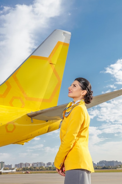 Fröhlicher Flugbegleiter, der den Himmel betrachtet und lächelt, während er in der Nähe eines gelben Flugzeugs am Flughafen steht