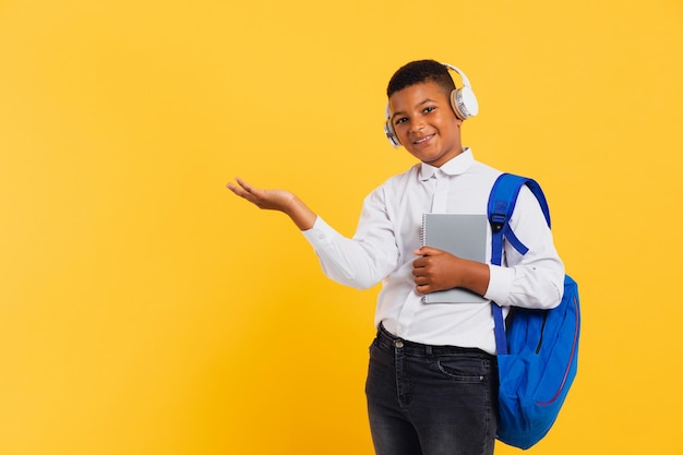 Fröhlicher gemischter Schuljunge mit Kopfhörern und Rucksack, der Bücher und Notizbücher hält und auf den Kopierbereich zeigt Zurück zum Schulkonzept