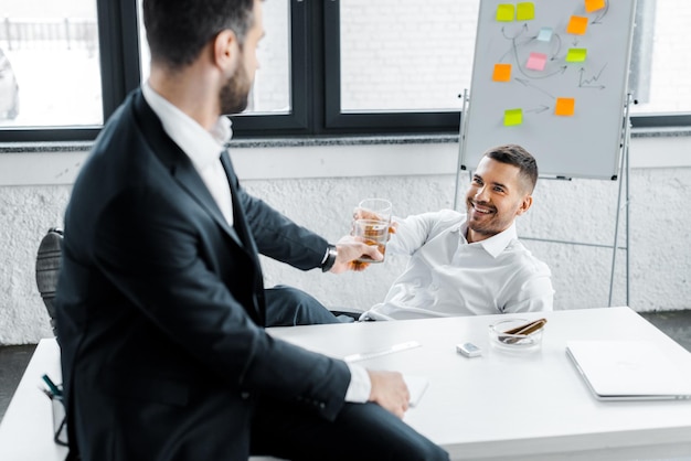 Fröhlicher Geschäftsmann, der mit einem Kollegen im modernen Büro mit Alkoholgetränk im Glas klirrt