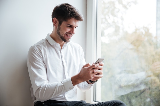 Fröhlicher Geschäftsmann in weißem Hemd, der im Café sitzt und beim Chatten auf das Telefon schaut