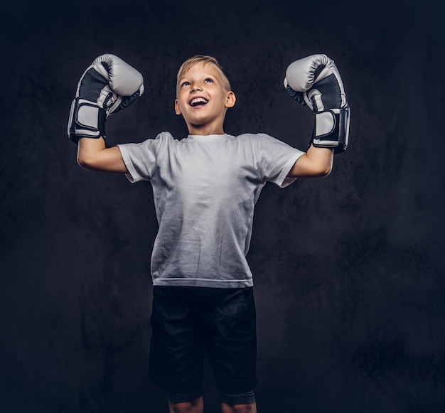 Foto fröhlicher, gutaussehender boxer des kleinen jungen mit blonden haaren, gekleidet in ein weißes t-shirt mit boxhandschuhen, freut sich über einen sieg. getrennt auf einem dunklen strukturierten hintergrund.