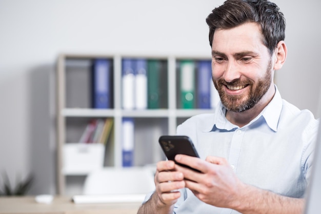 Fröhlicher gutaussehender Büroangestellter, der mit einem Mobiltelefon an einem Computertisch im Büro sitzt Fröhlicher bärtiger Geschäftsmann Unternehmer Mitarbeiter durchsucht Smartphone liest Nachrichten Chats kommuniziert