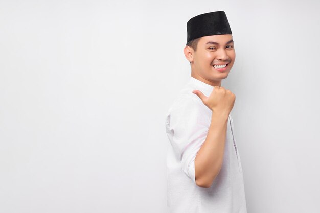 Fröhlicher, gutaussehender junger asiatischer muslimischer Mann in arabischem Kostüm, der mit dem Finger auf einen freien Platz für eine Werbung zeigt, die auf weißem Hintergrund isoliert ist Menschen religiöses islamisches Lebensstilkonzept